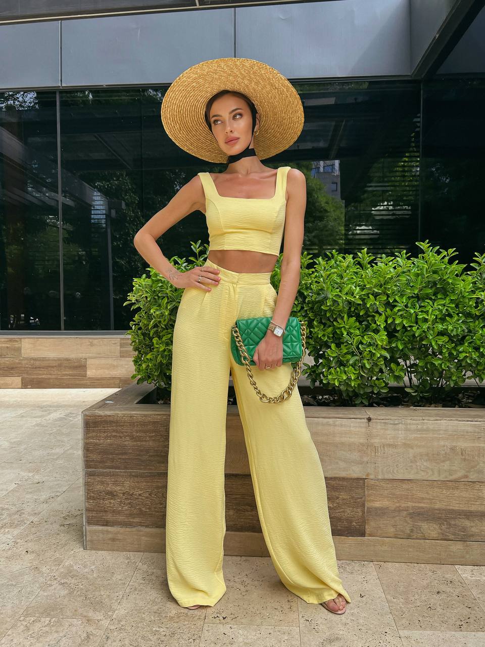 Woman in yellow outfit with wide-brimmed hat and green clutch standing outdoors.