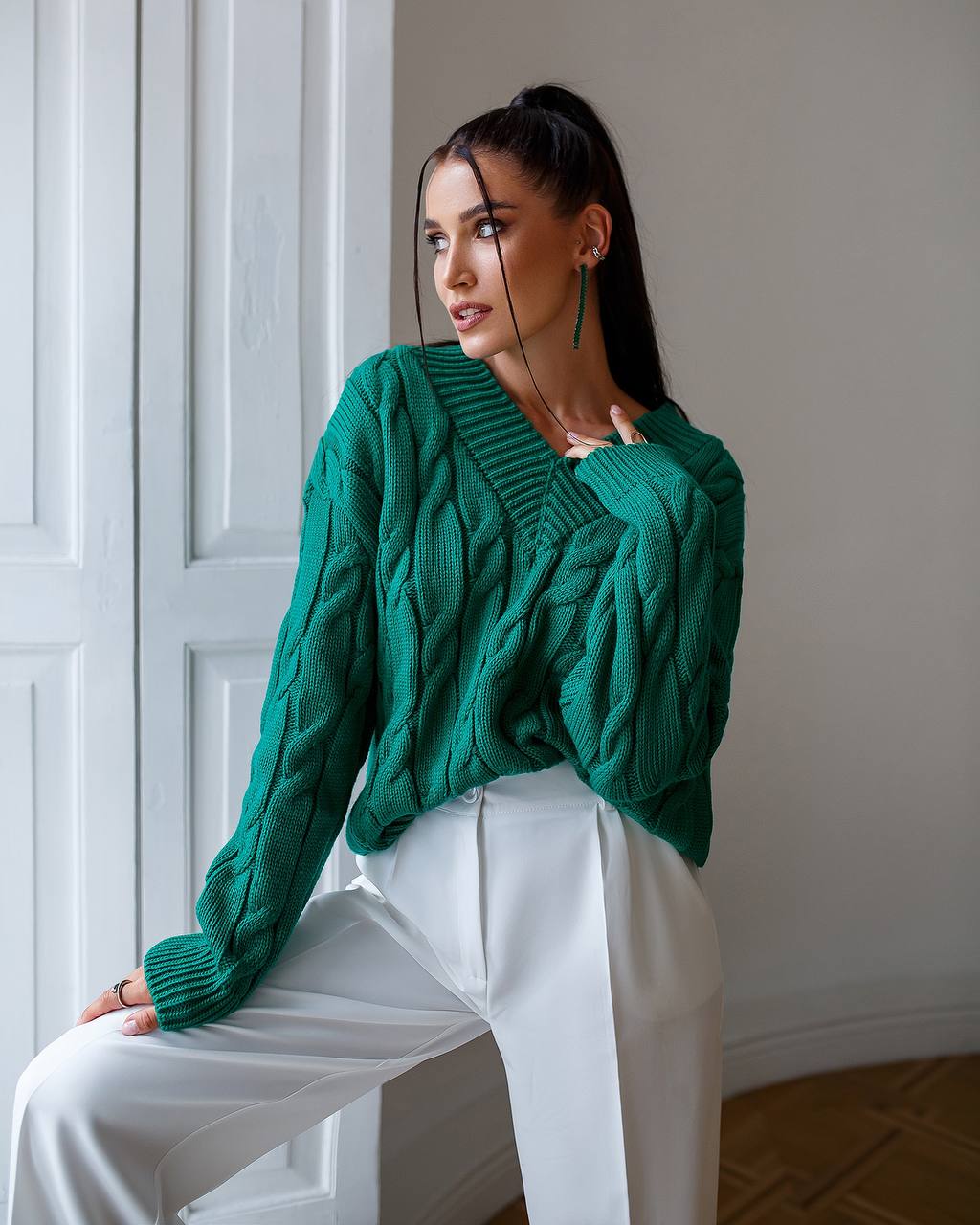 Woman wearing a green sweater and white pants sitting indoors.