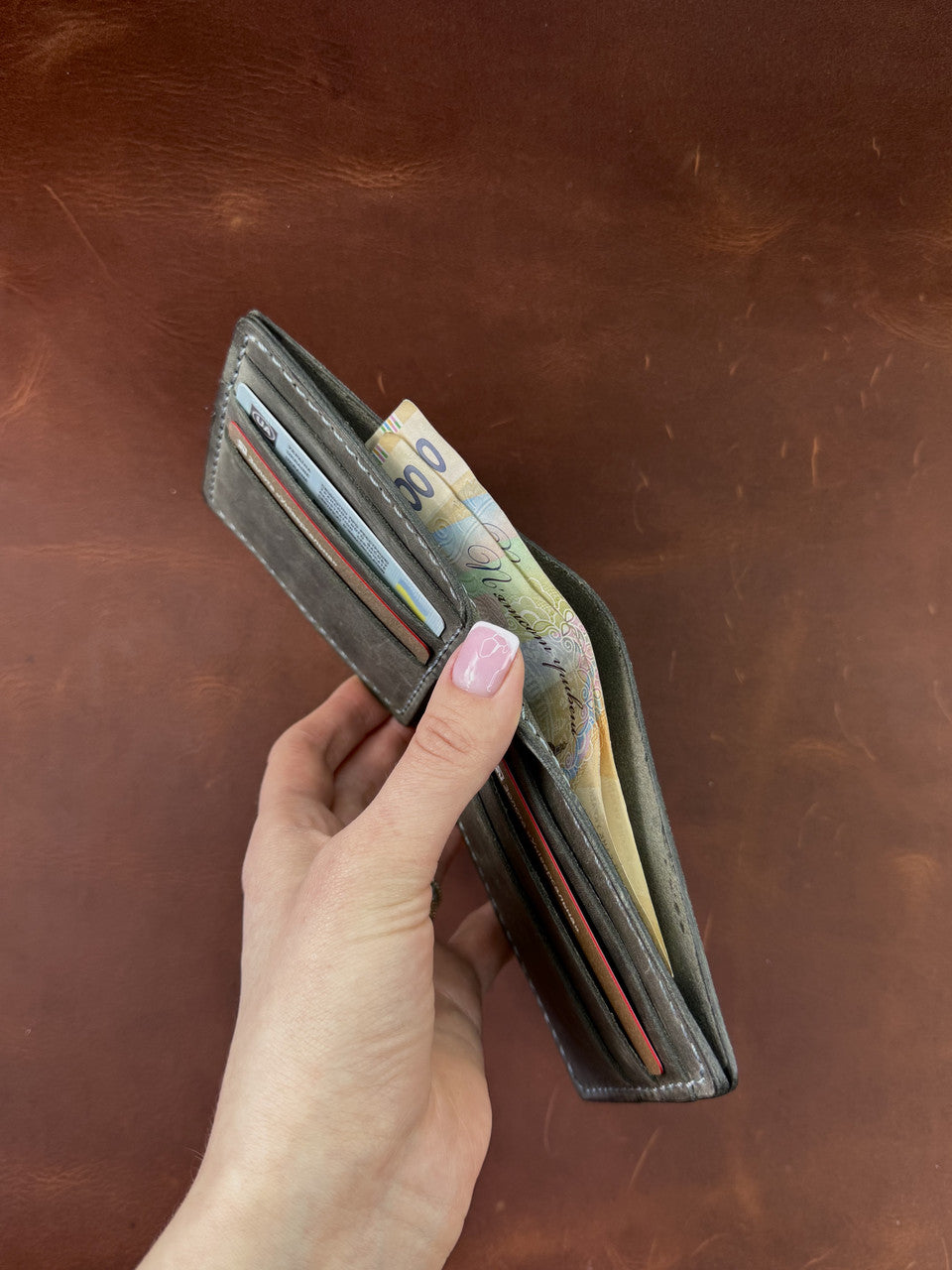 A hand with pink nail polish holds an open dark brown leather bifold wallet containing cards and Australian dollar banknotes, set against a matching brown leather background.
