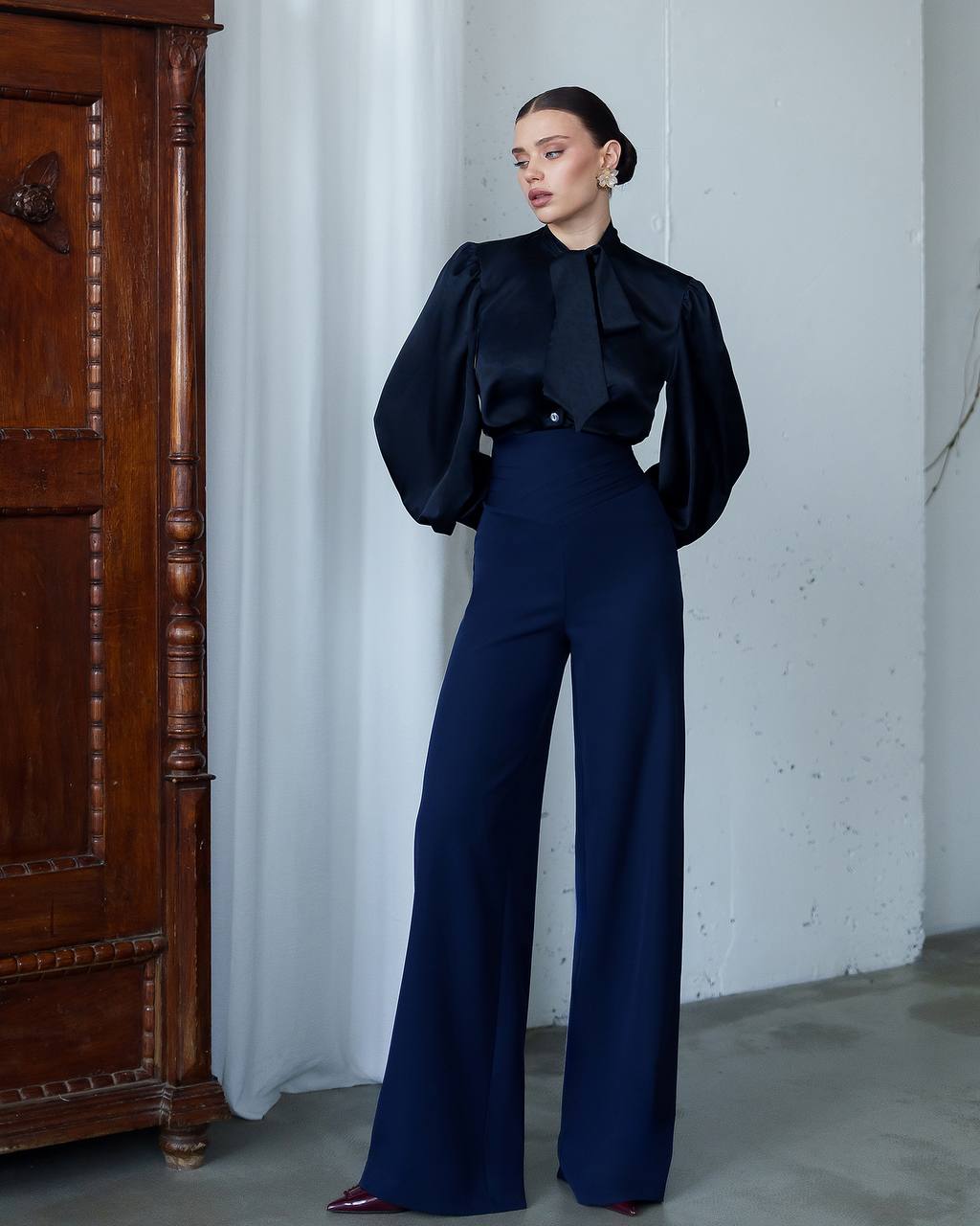 Woman in a black top and blue pants standing against a white wall with a wooden cabinet.