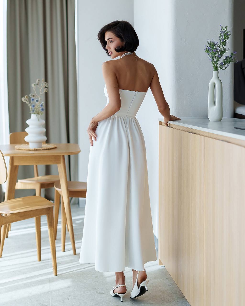 Woman in a white strapless dress standing in a modern room with wooden furniture and decor.