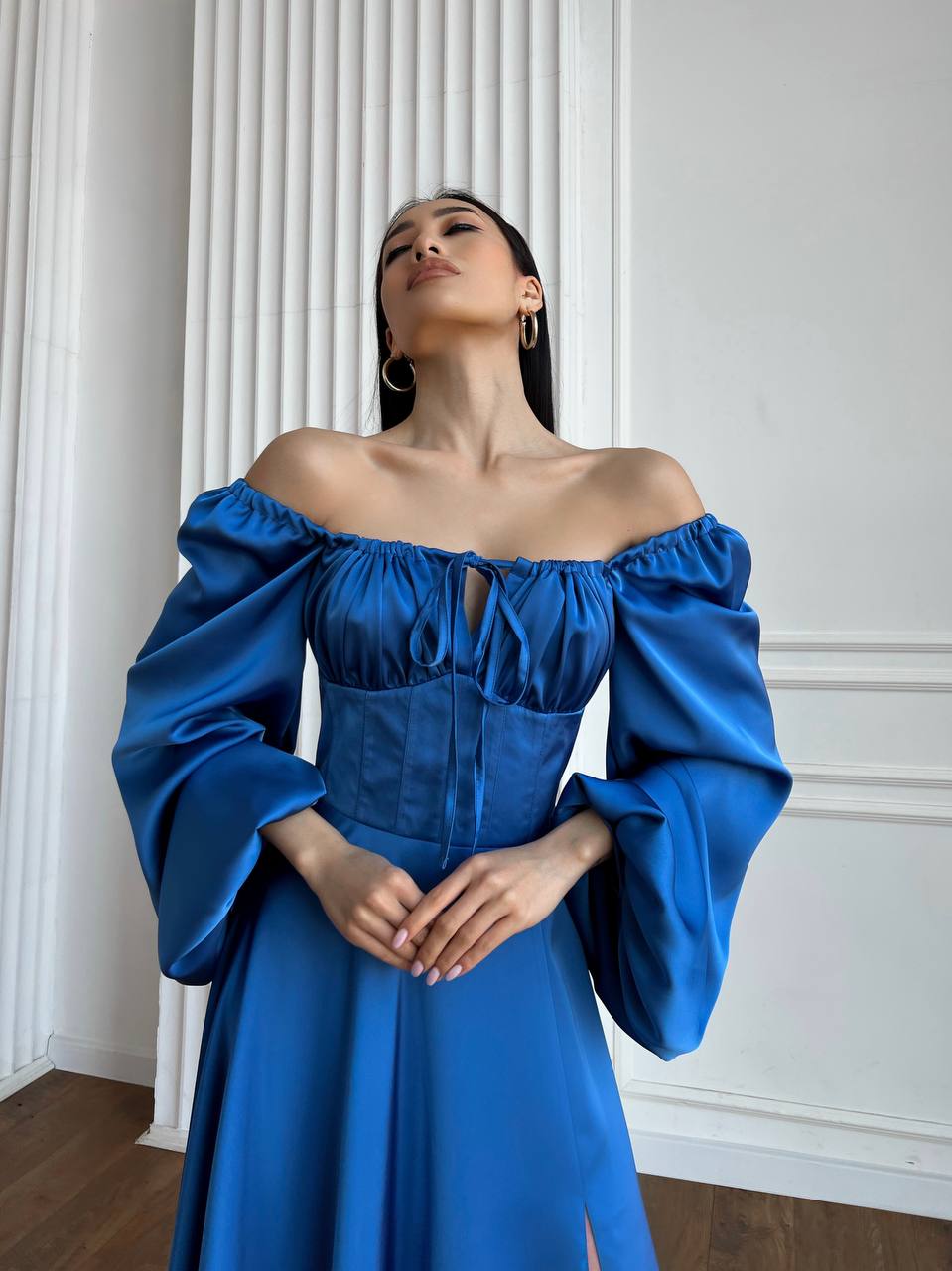 Woman wearing a blue off-shoulder dress against a white paneled wall.