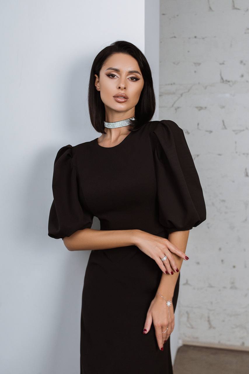 Woman wearing a black dress with puffed sleeves against a light gray wall.