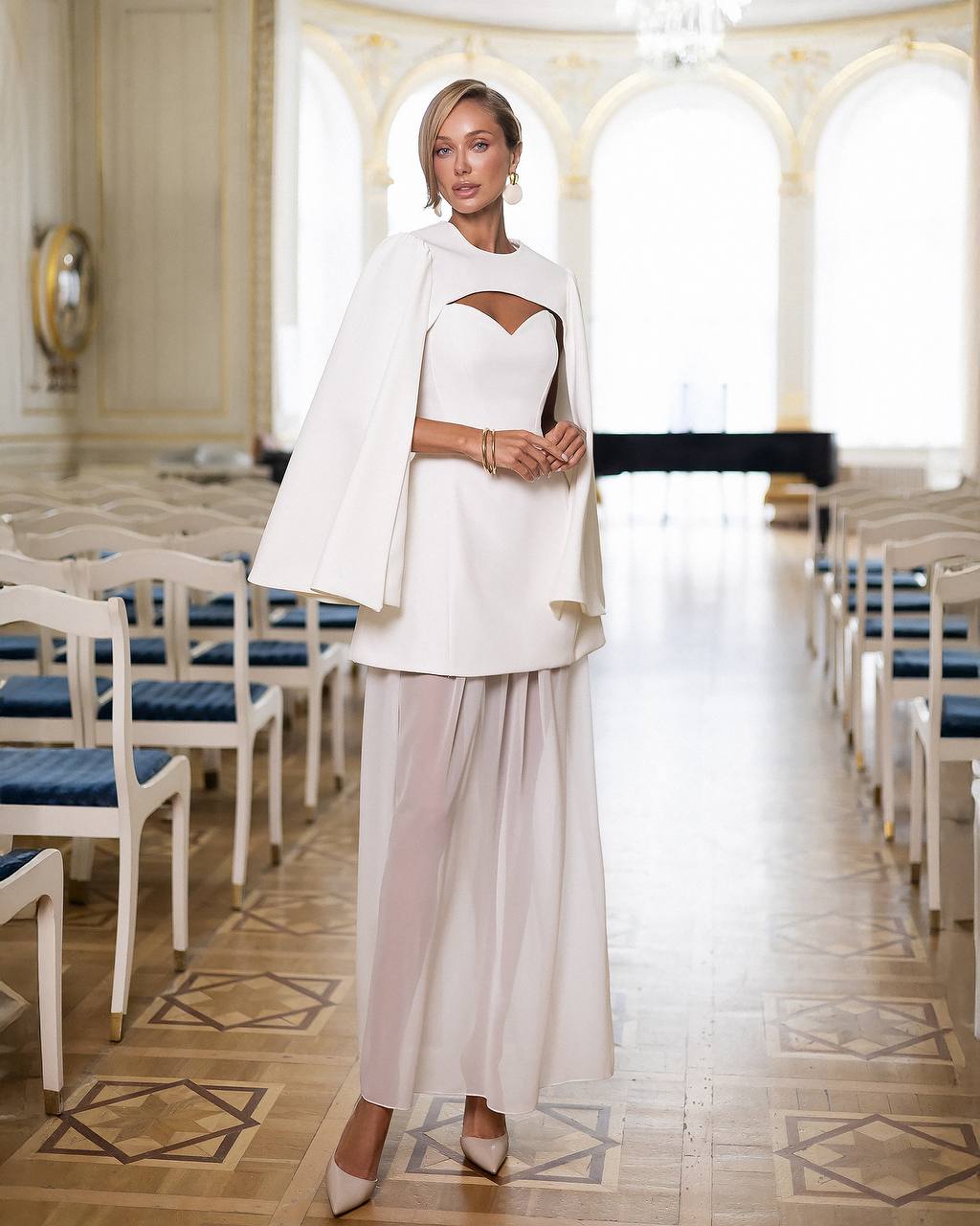 Woman in a white outfit standing in an elegant indoor setting with large windows.