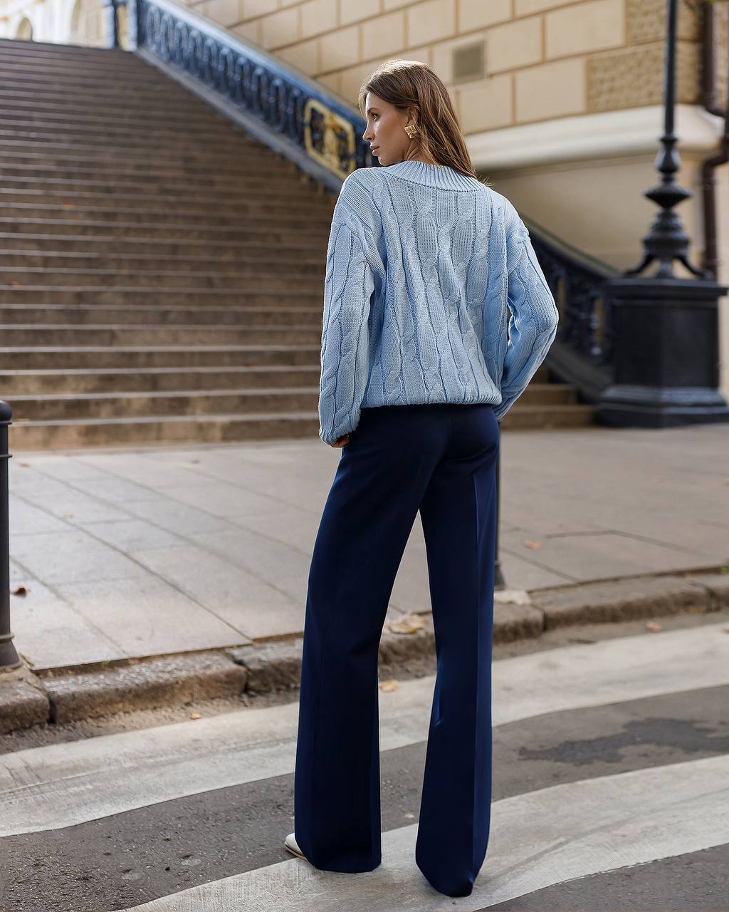 Woman wearing a light blue cable knit sweater and dark blue pants on a city street.