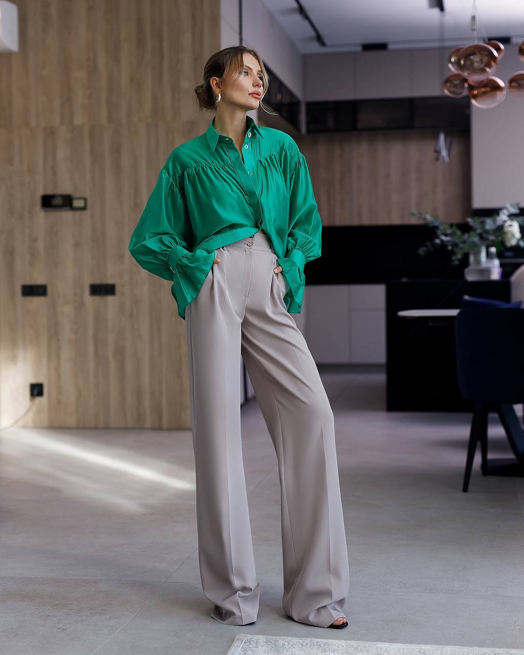Woman in a green blouse and beige pants standing in a modern indoor setting.