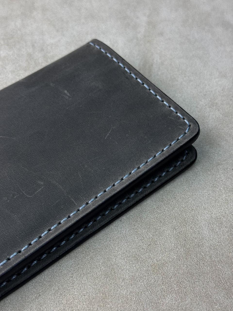 A close-up of a black leather wallet or card holder with light blue stitching, placed on a textured gray surface.