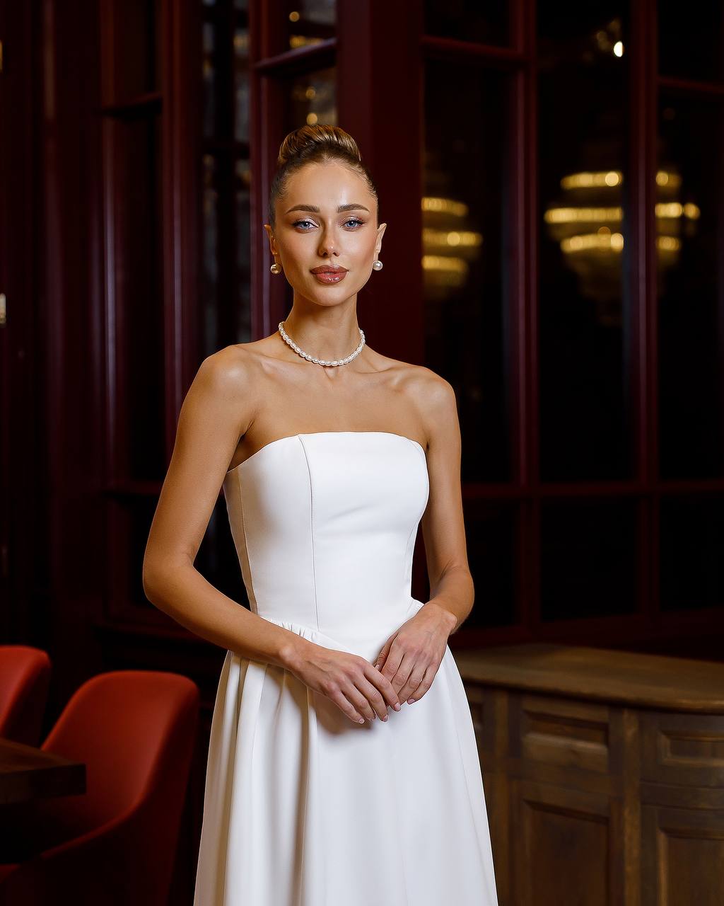 Woman in a white strapless dress standing in an indoor setting with red chairs and dark wood paneling.