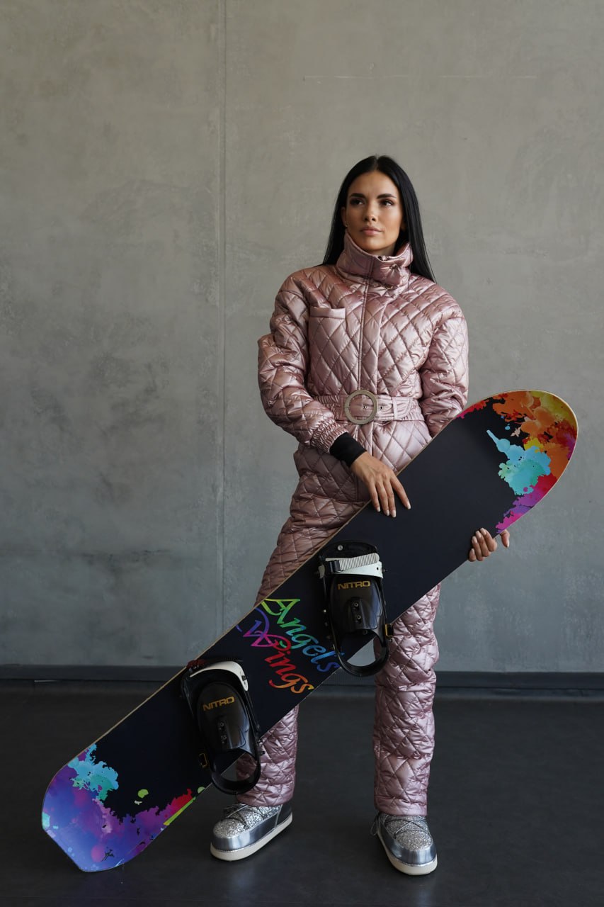 Person in a pink quilted jacket and pants holding a colorful snowboard against a gray wall.