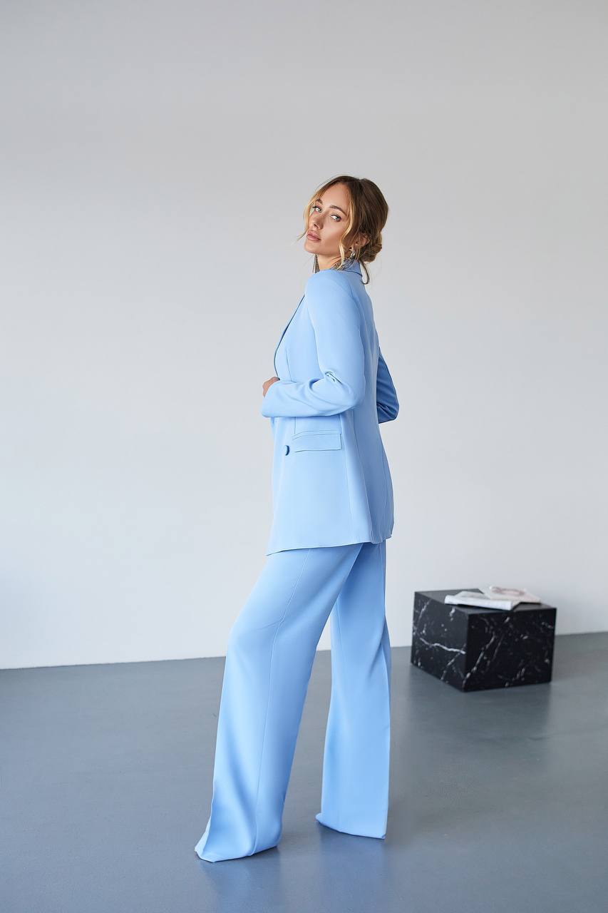 Woman wearing a light blue suit standing in a minimalistic room with a white wall and gray floor.