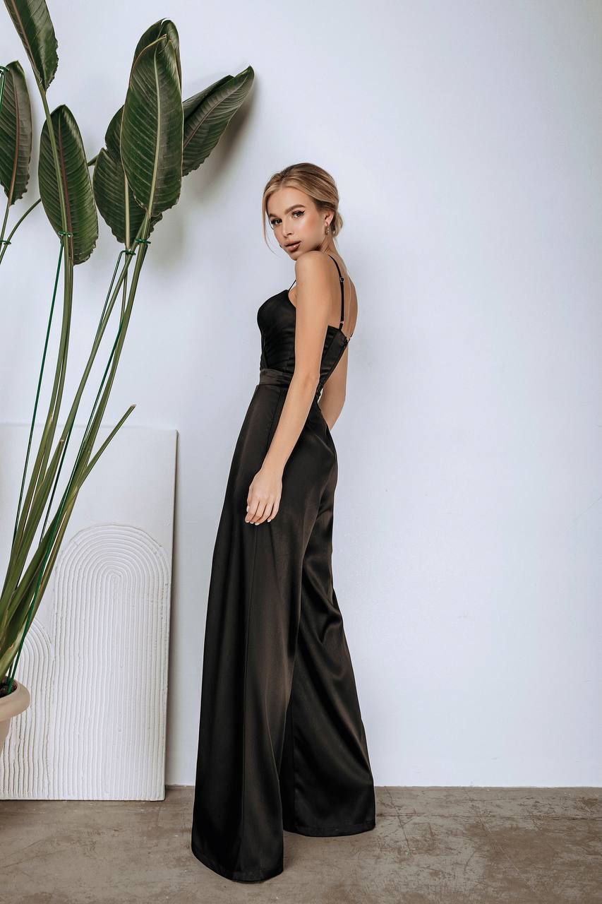 Woman in a black evening gown standing next to a large plant against a white wall.