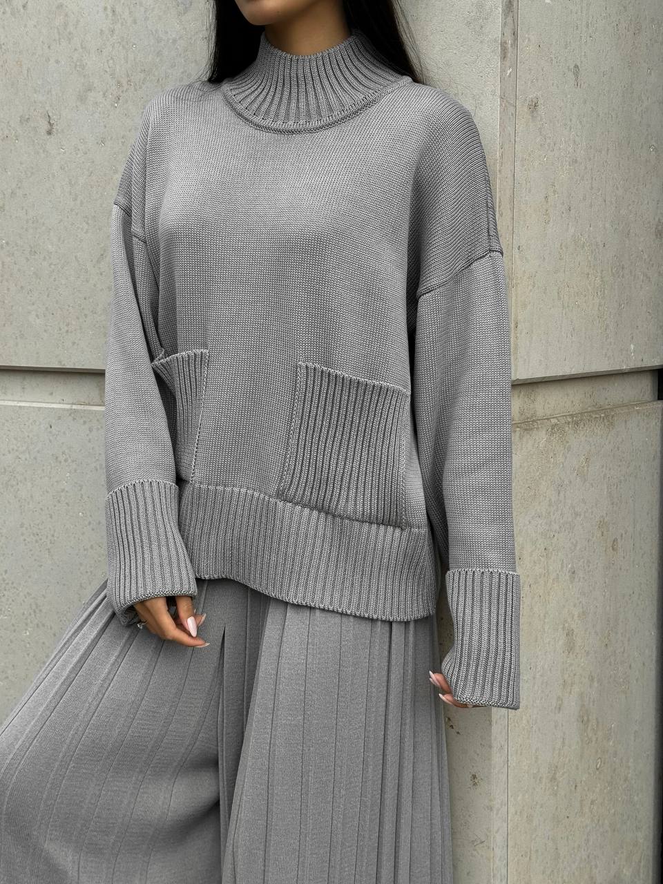 Person wearing a gray ribbed sweater and pants against a concrete wall.