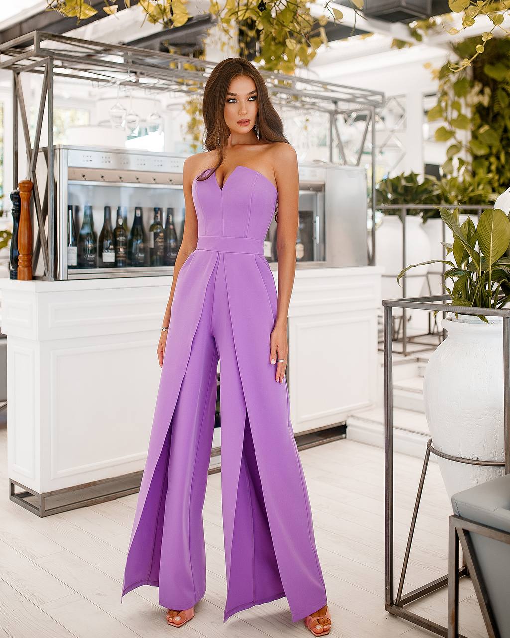 Woman wearing a purple jumpsuit in an indoor setting with plants and furniture.