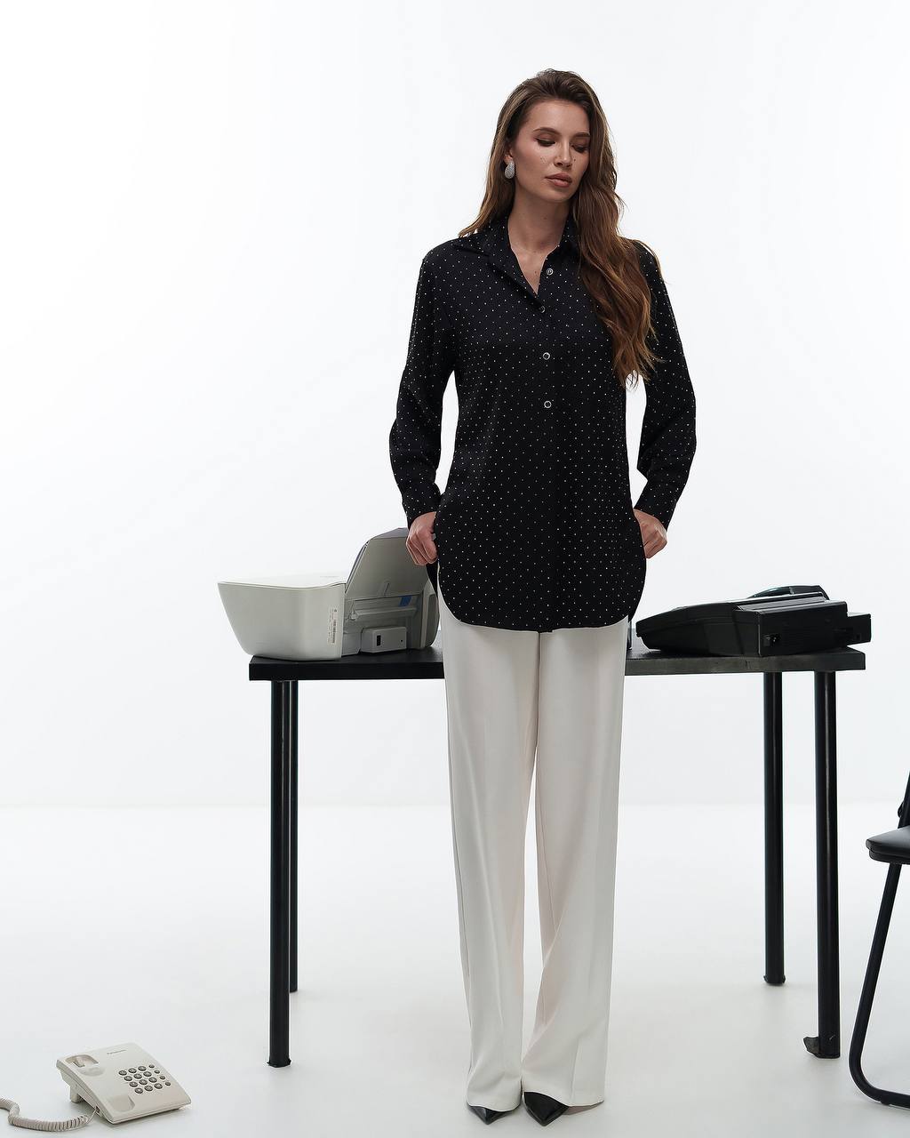 Woman in a black polka dot blouse and white pants standing in front of a desk with office items.