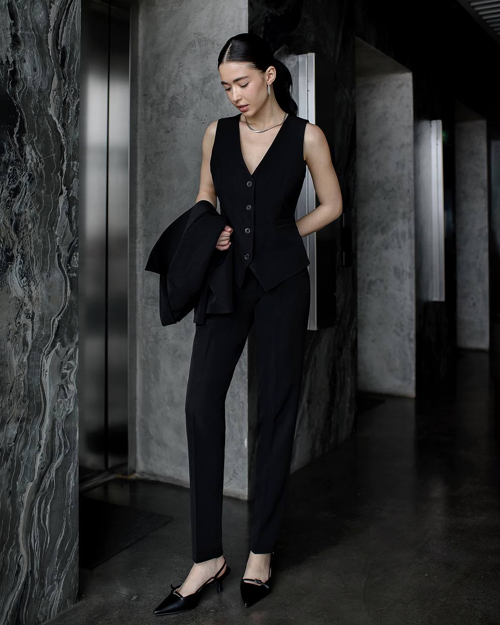 Woman in a black suit standing in a dimly lit room with marble walls.