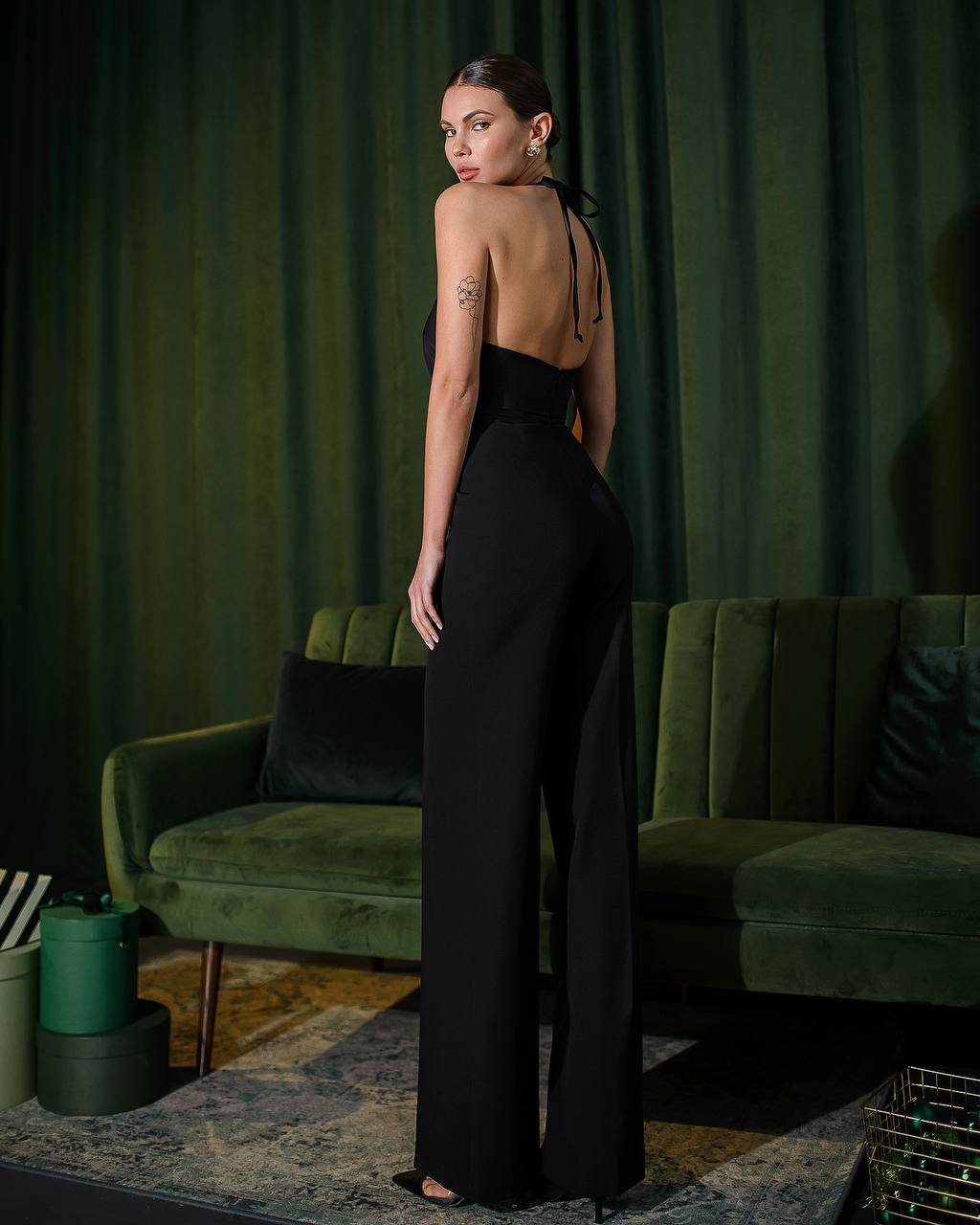 Woman in a black jumpsuit standing in a room with green curtains and a green couch.