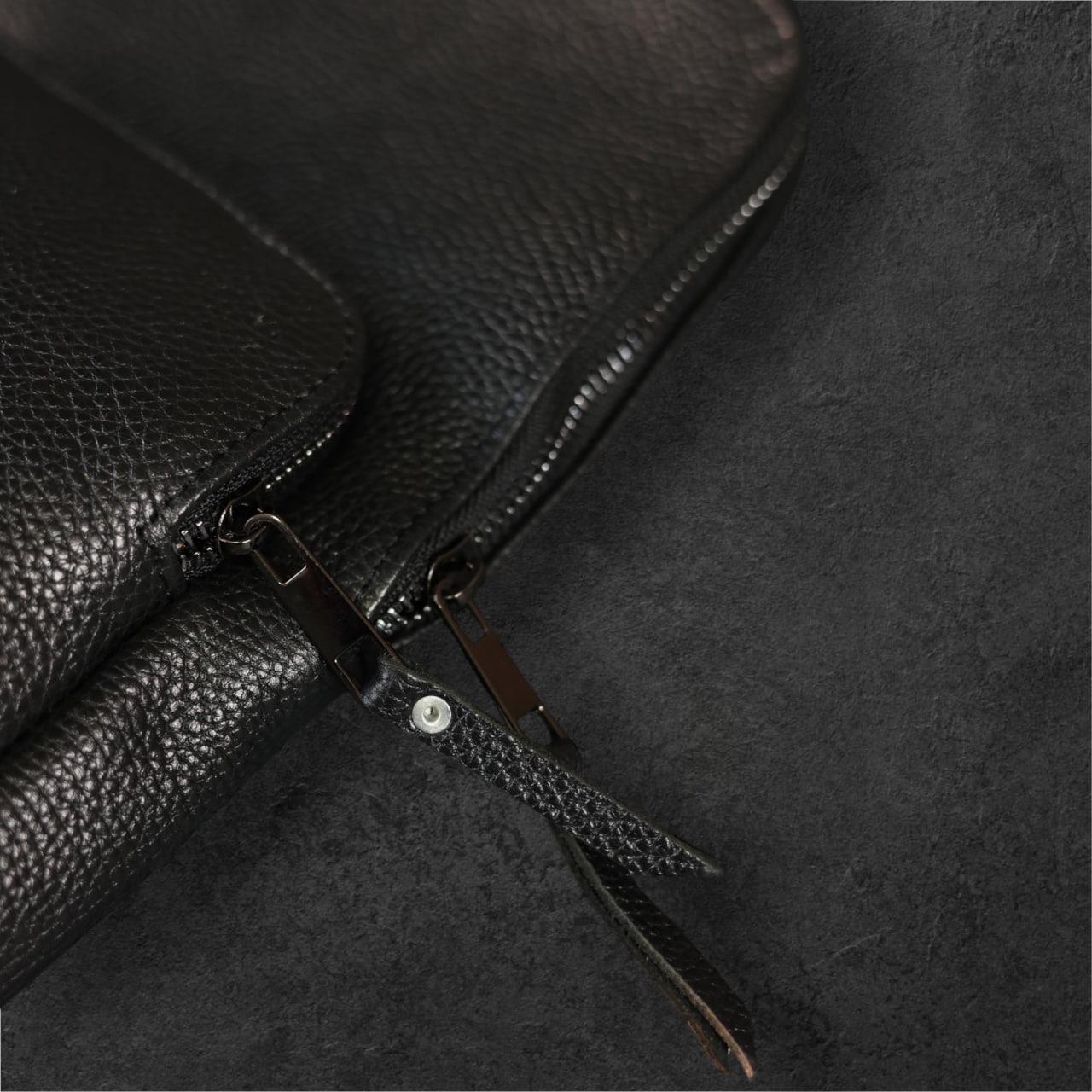 Close-up of a black pebbled leather bag with a zipper and leather pull tab on a dark textured background.