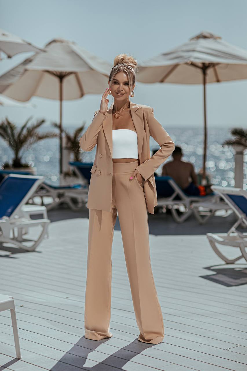 Woman in a beige suit standing on a beach deck with umbrellas and lounge chairs in the background.
