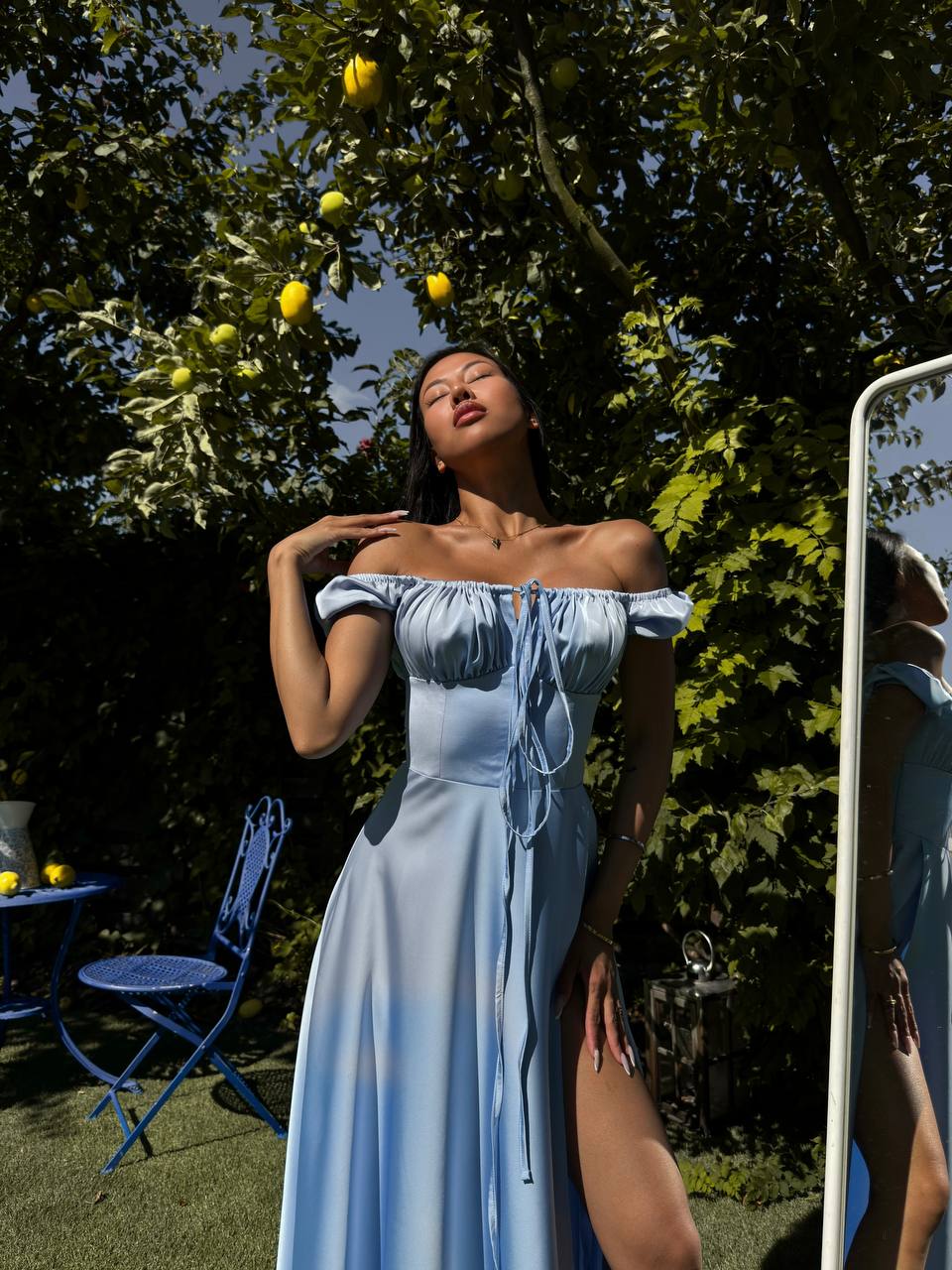 Woman in a light blue dress standing outdoors with trees and a mirror in the background