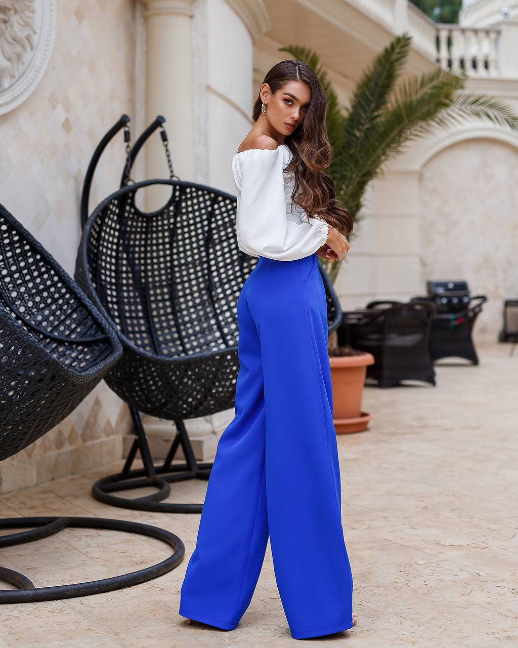 Woman in white top and blue pants standing in an outdoor setting with chairs and plants.
