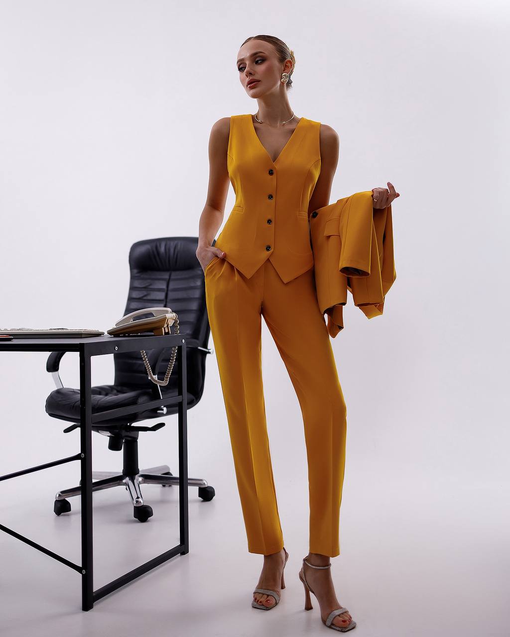 Woman in a mustard yellow suit standing next to a black office chair on a white background