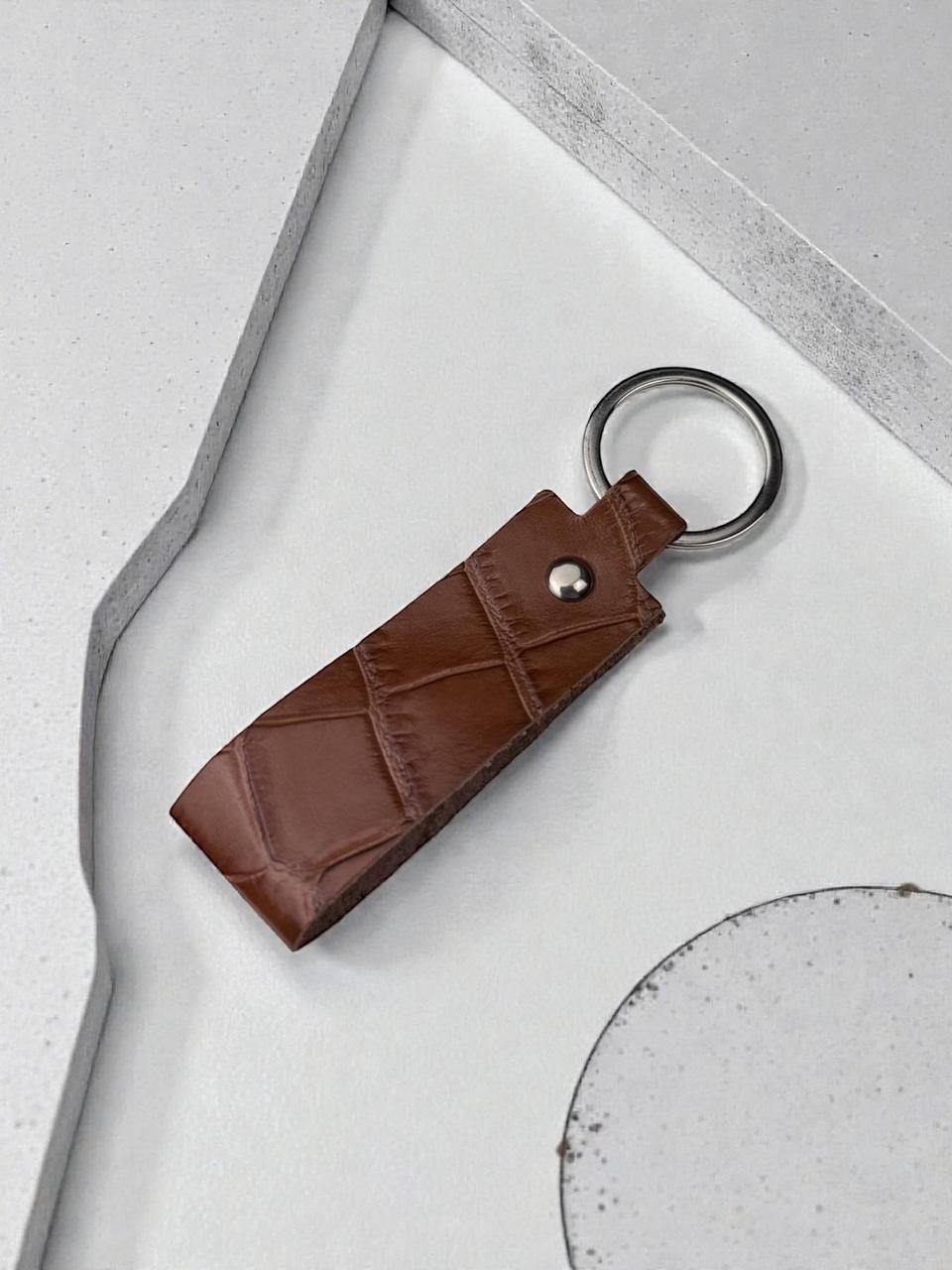 A brown crocodile-embossed leather keychain with a silver metal key ring and rivet, displayed on a modern, textured light gray concrete surface.