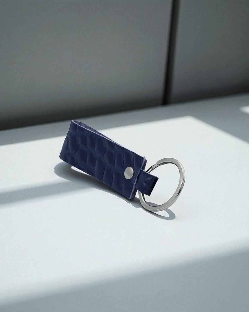 A dark blue crocodile-embossed leather keychain with a silver metal split ring, resting on a light gray surface with soft shadows.
