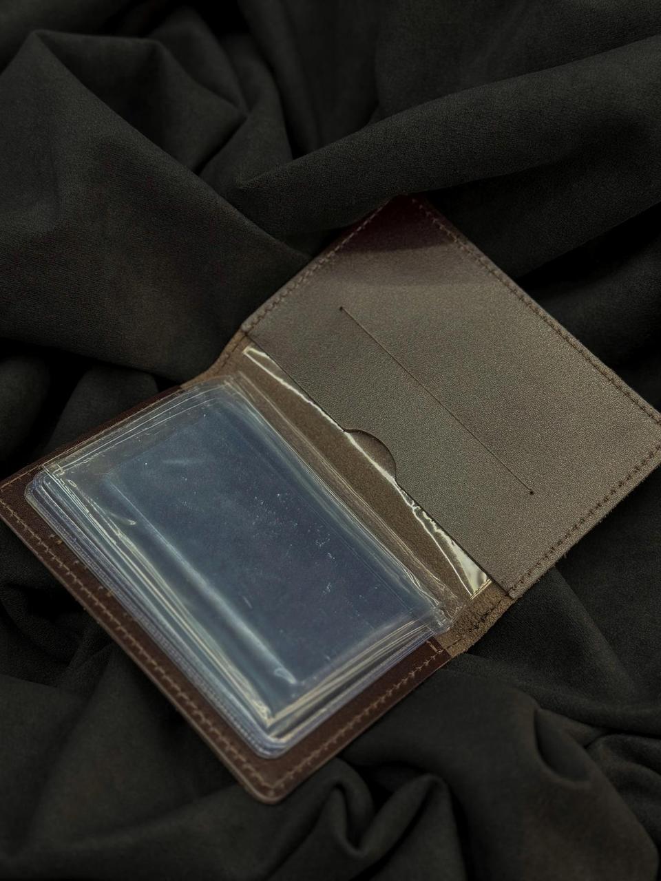 A brown leather card holder wallet shown open, featuring a clear plastic ID window on the front, resting on a dark charcoal gray fabric background.