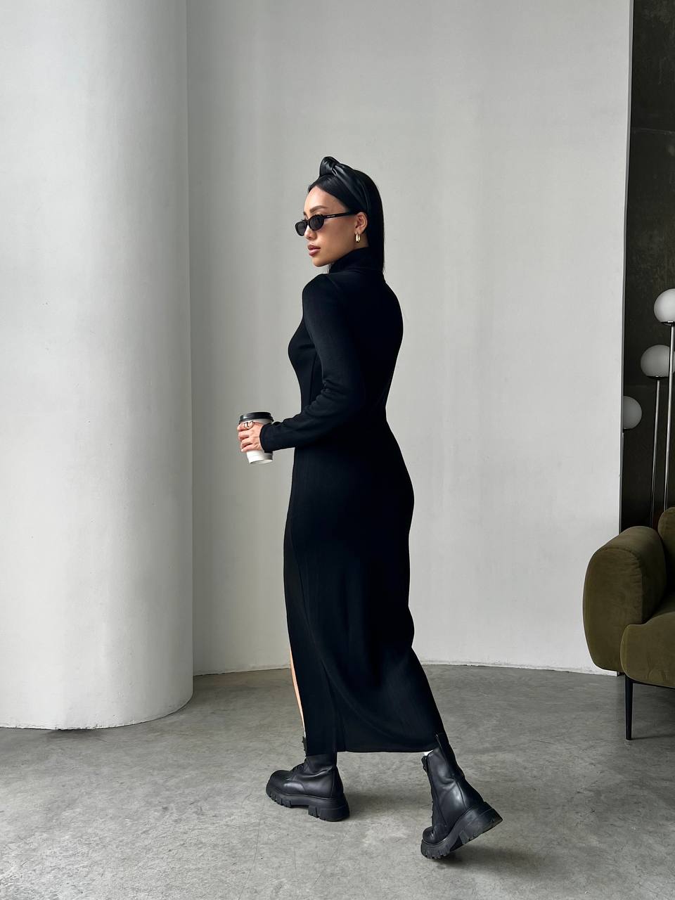 Person wearing a long black dress and black shoes, holding a coffee cup, in a minimalistic room.