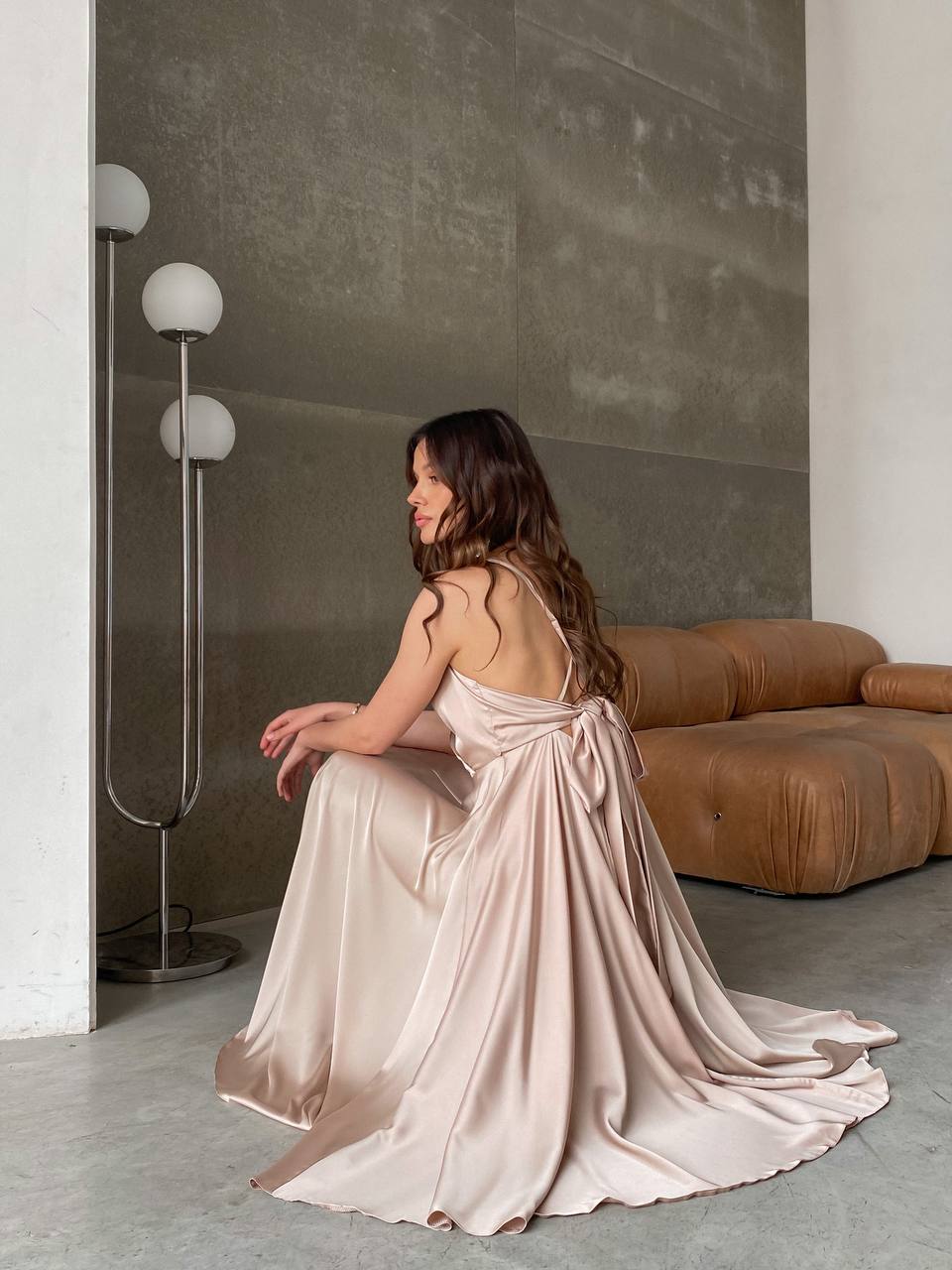 Woman in a flowing dress sitting on the floor in a modern room with a brown sofa and lamp.