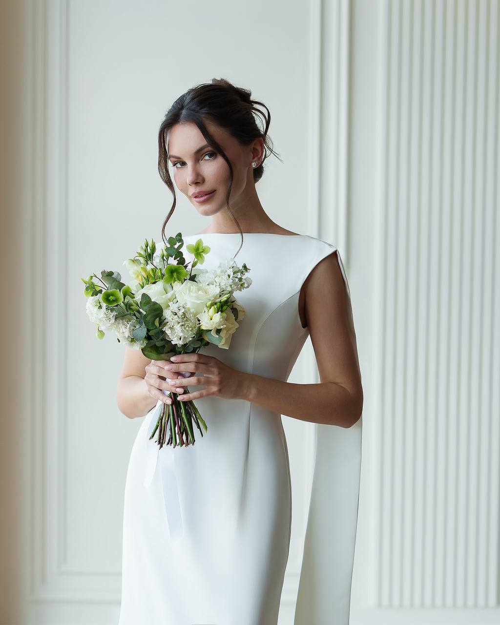 Woman in a white dress holding a bouquet of flowers against a white wall.