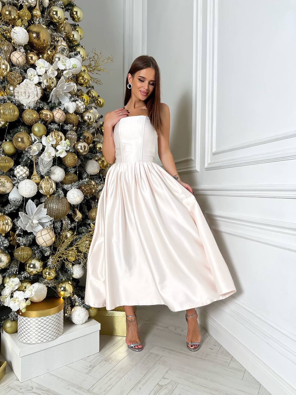 Woman in a white dress standing next to a decorated Christmas tree in a room with white walls.