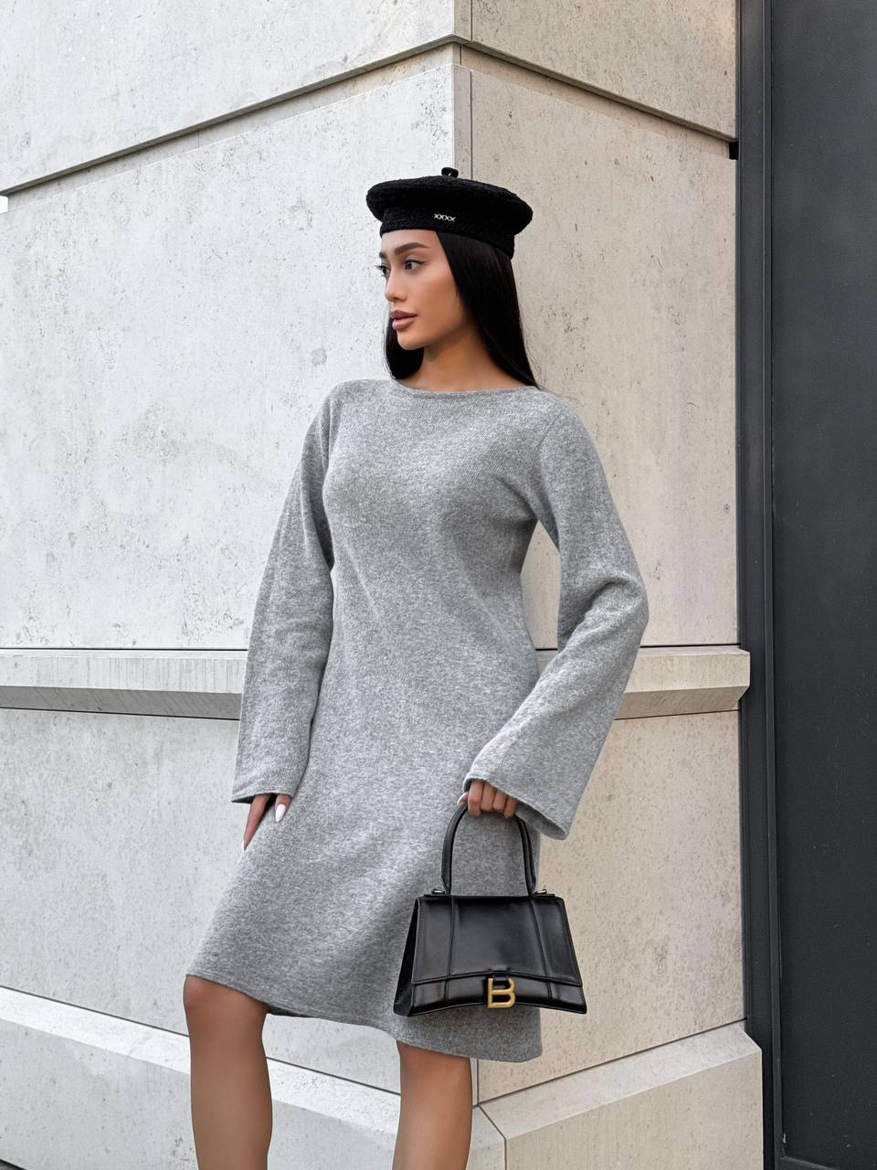 Woman in a gray dress and black hat holding a black handbag against a light-colored wall.