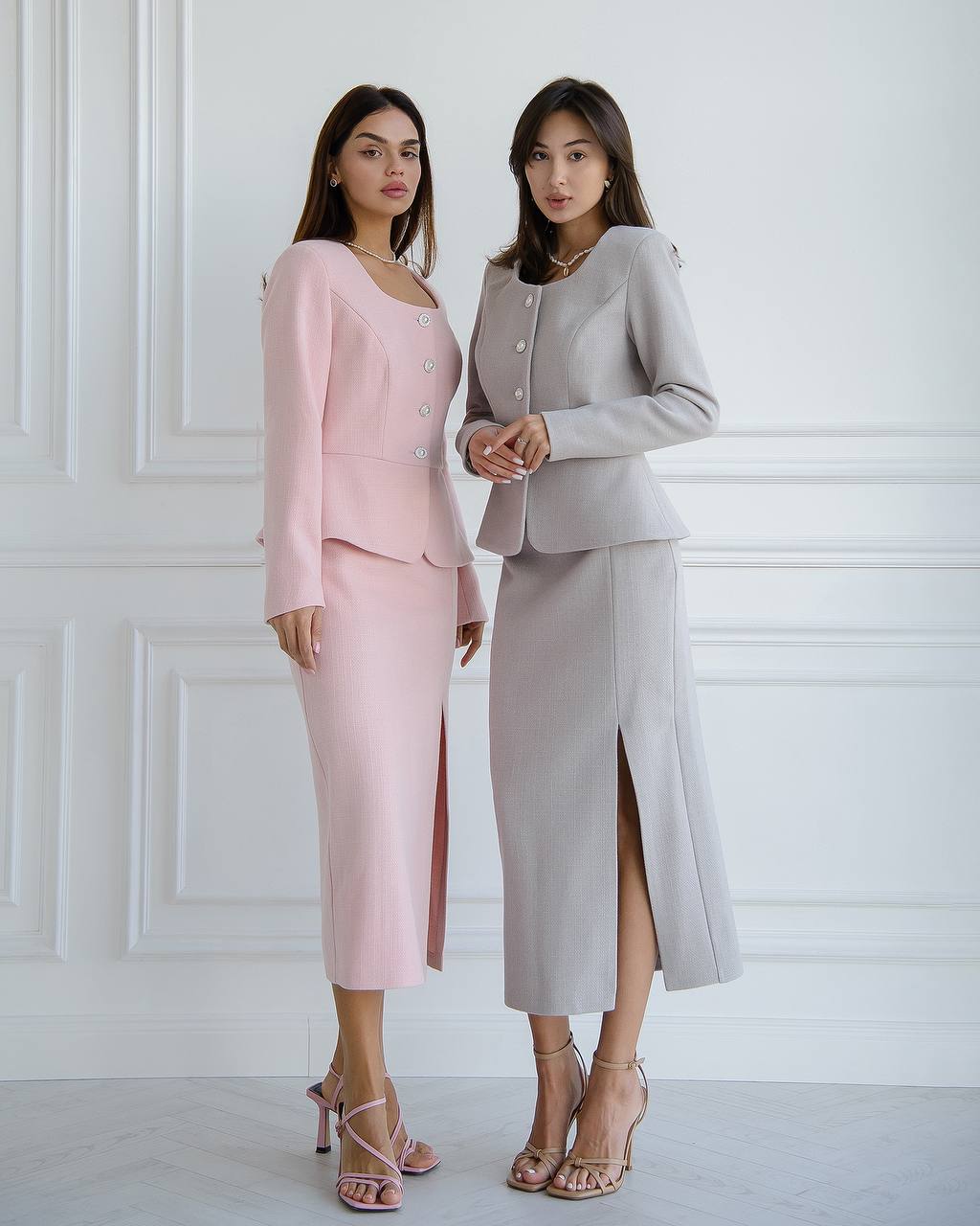 Two women wearing matching pink and gray suits against a white wall.