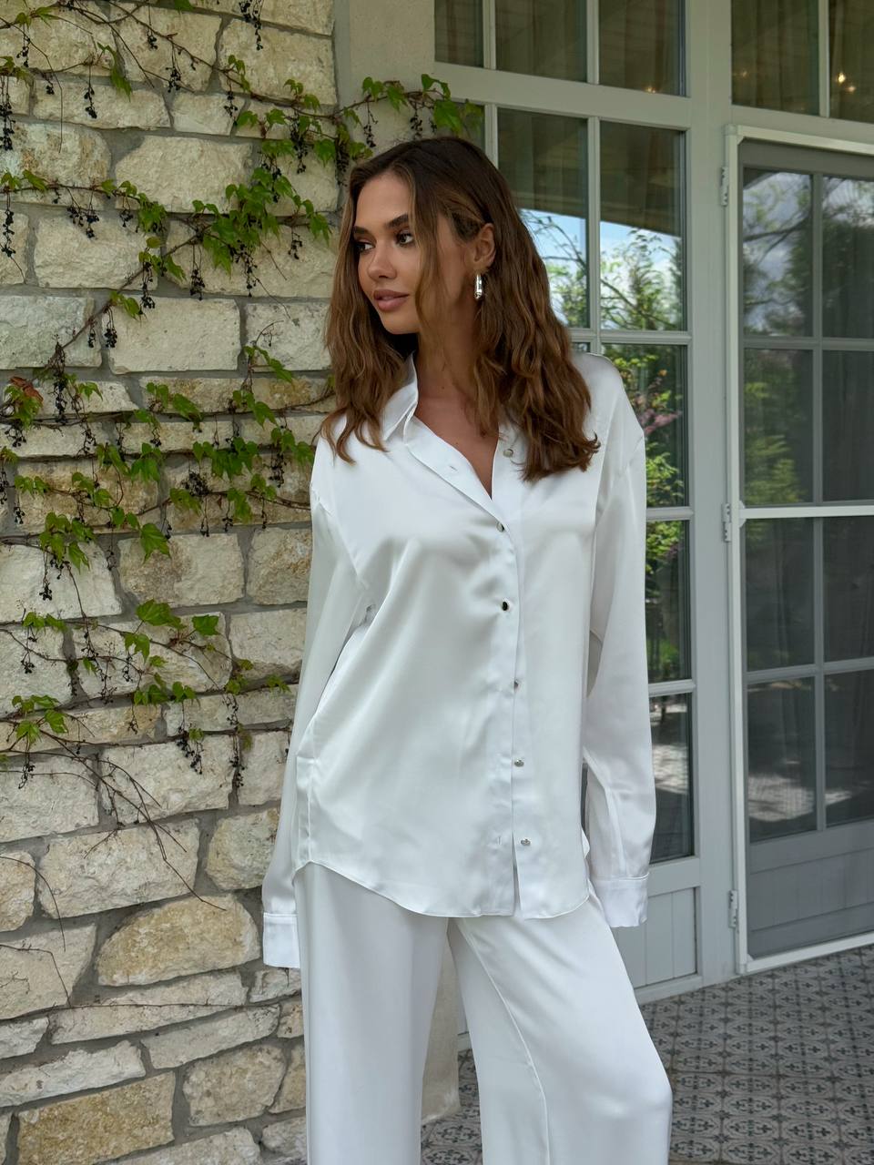 Woman in a white outfit standing against a stone wall with greenery.