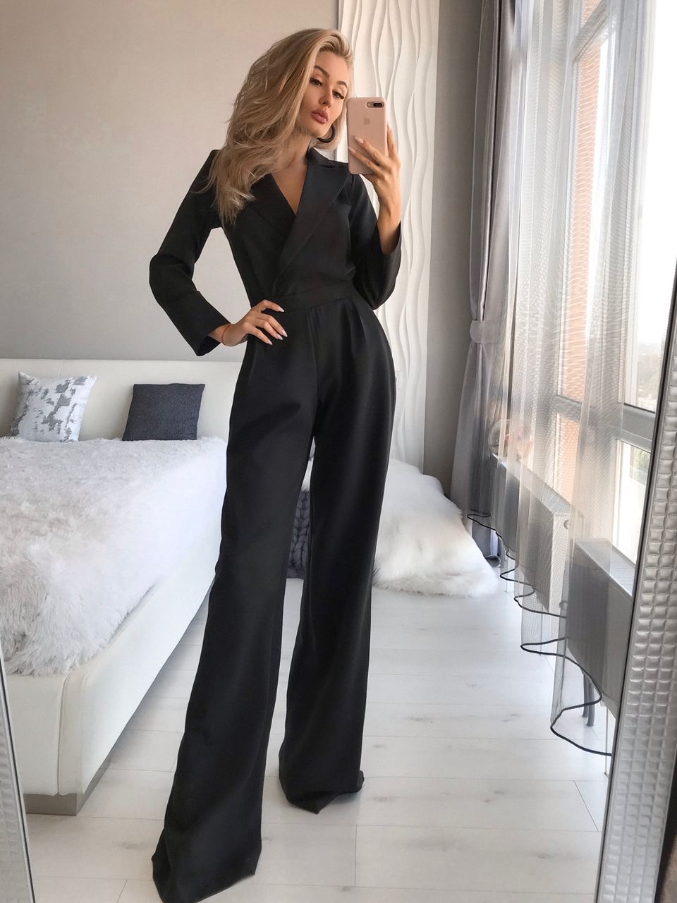 Woman in a black jumpsuit taking a mirror selfie in a modern bedroom.