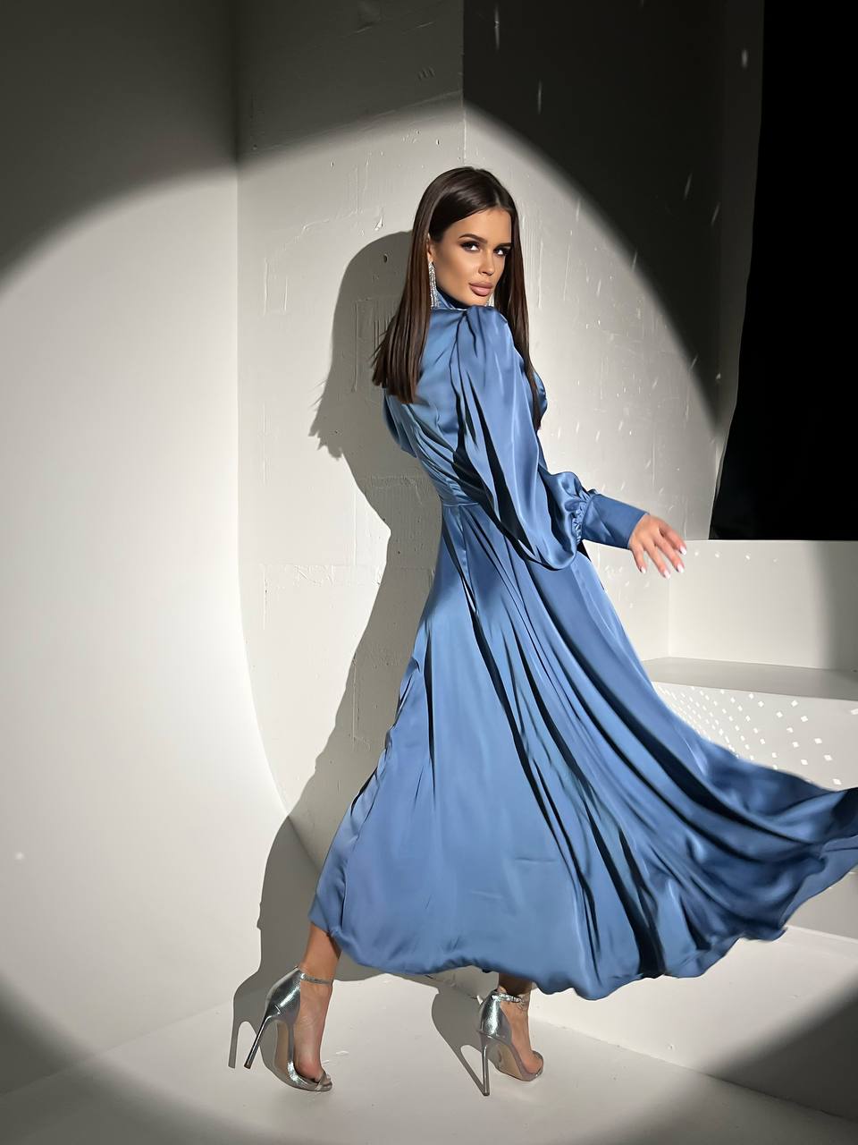 Woman in a flowing blue dress standing against a white wall with a spotlight effect.