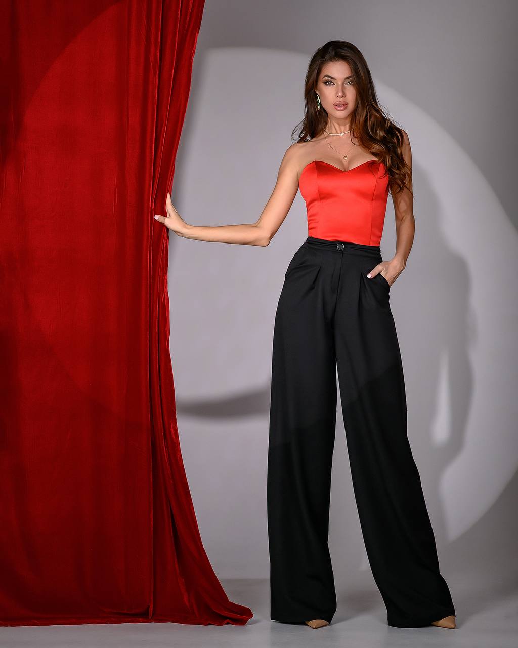 Woman in a red top and black pants standing next to a red curtain on a gray background