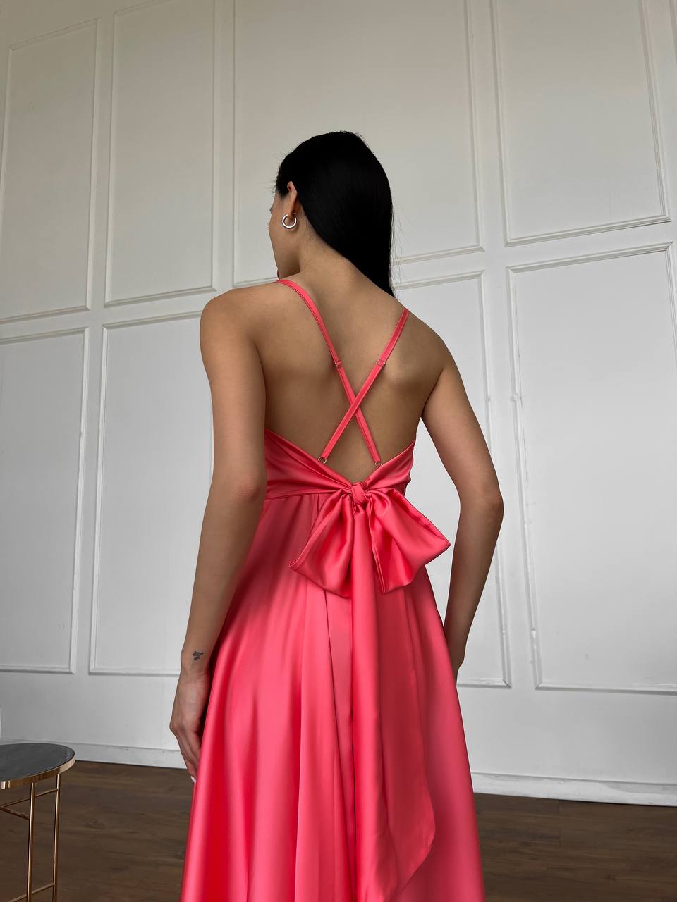 Woman wearing a pink dress with a bow detail in front of a white paneled wall.