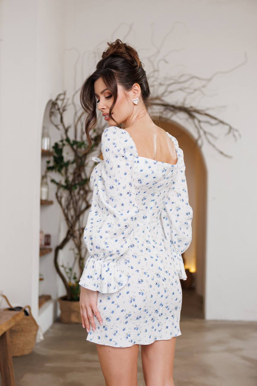 Woman wearing a white floral dress in an indoor setting with plants and a mirror.