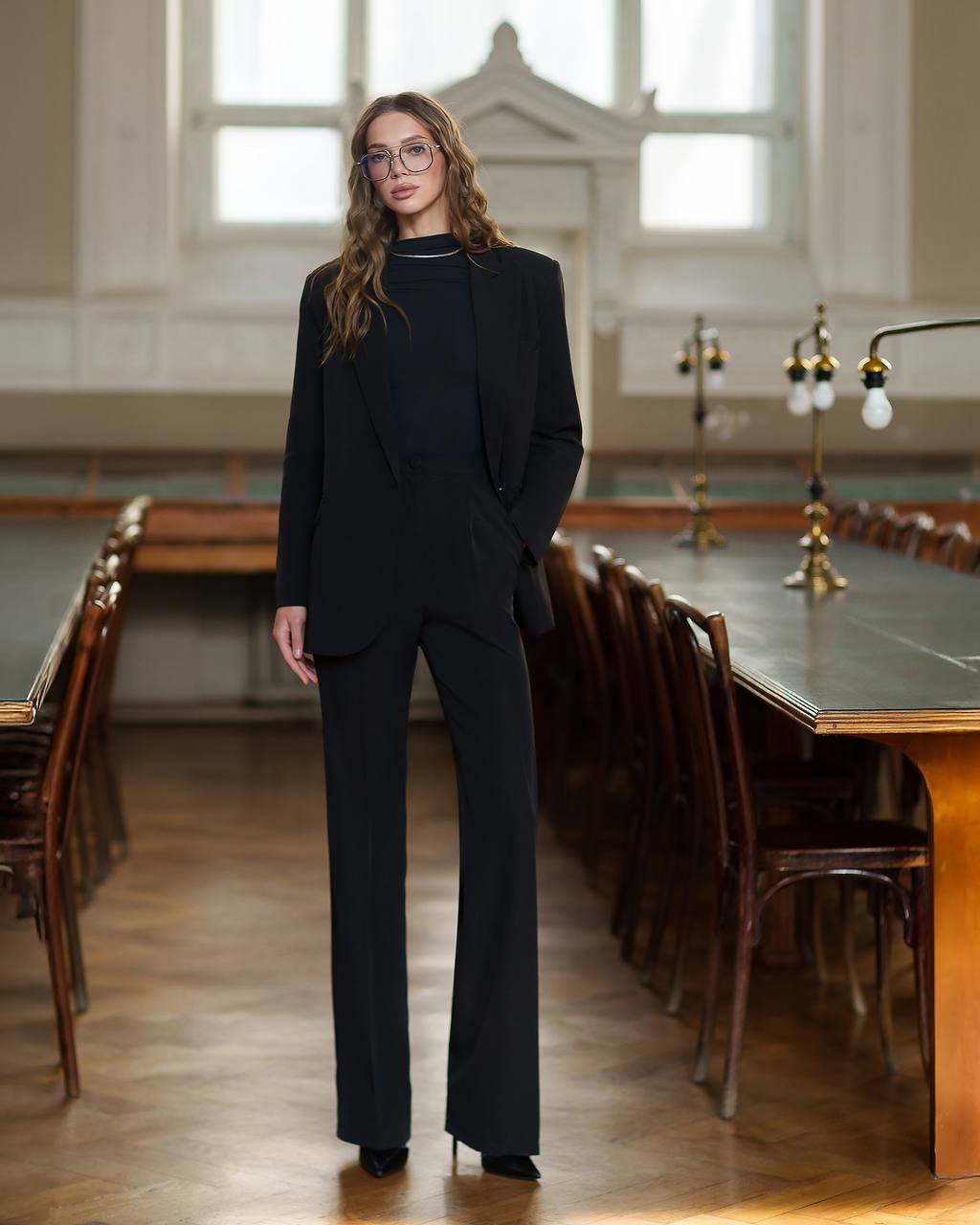 Woman in a black suit standing in a large room with wooden tables and chairs.