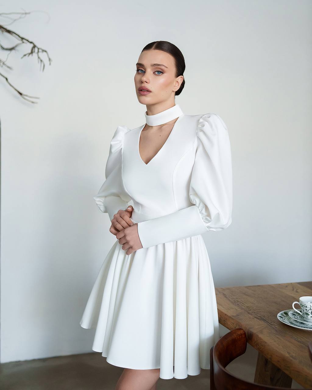 Woman wearing a white dress with long sleeves and a high neckline, standing in a room with a wooden table and decorative elements.