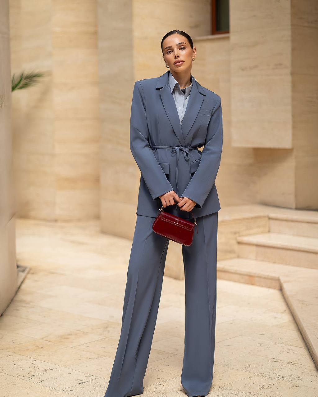 Woman in a gray suit holding a red handbag in front of a beige building.