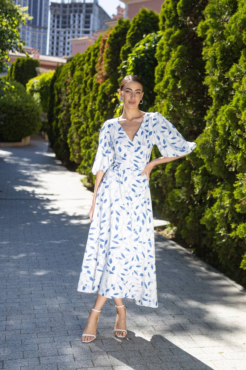 Woman in a white dress with blue patterns standing on a sidewalk with greenery in the background