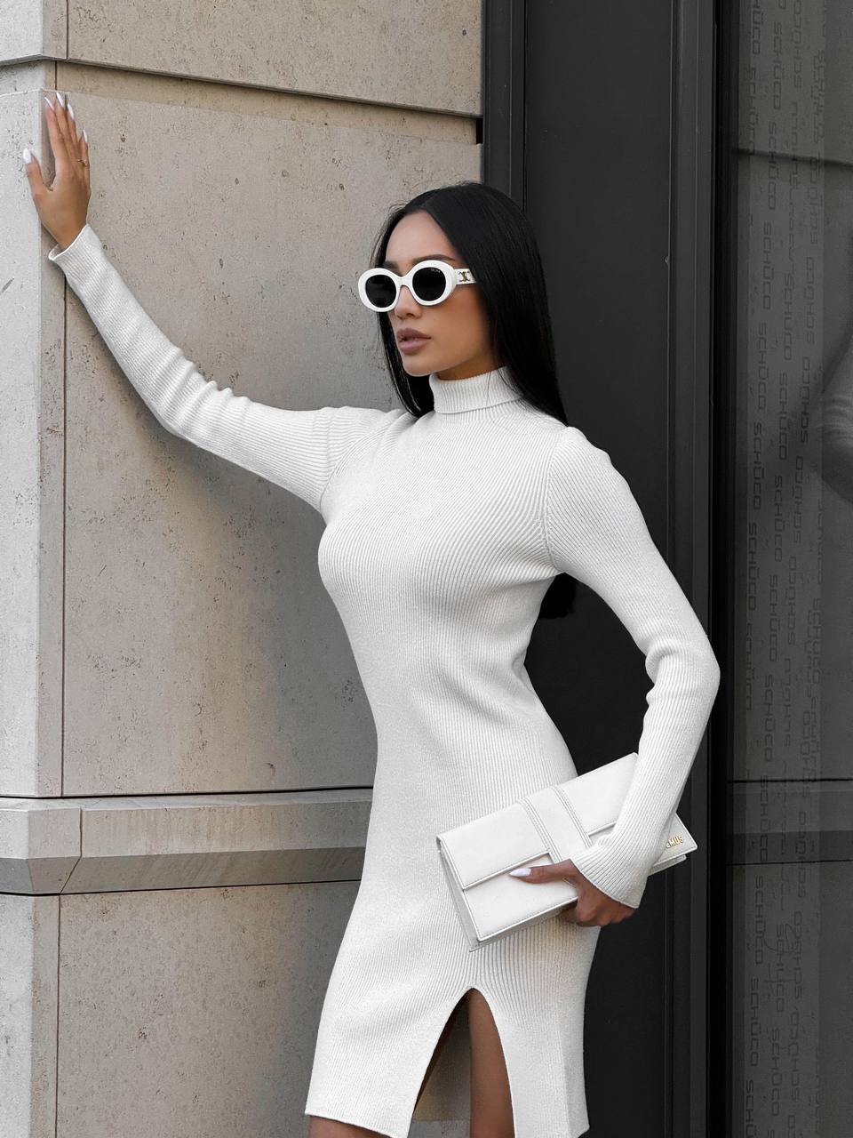Woman in a white dress and sunglasses standing against a building wall.