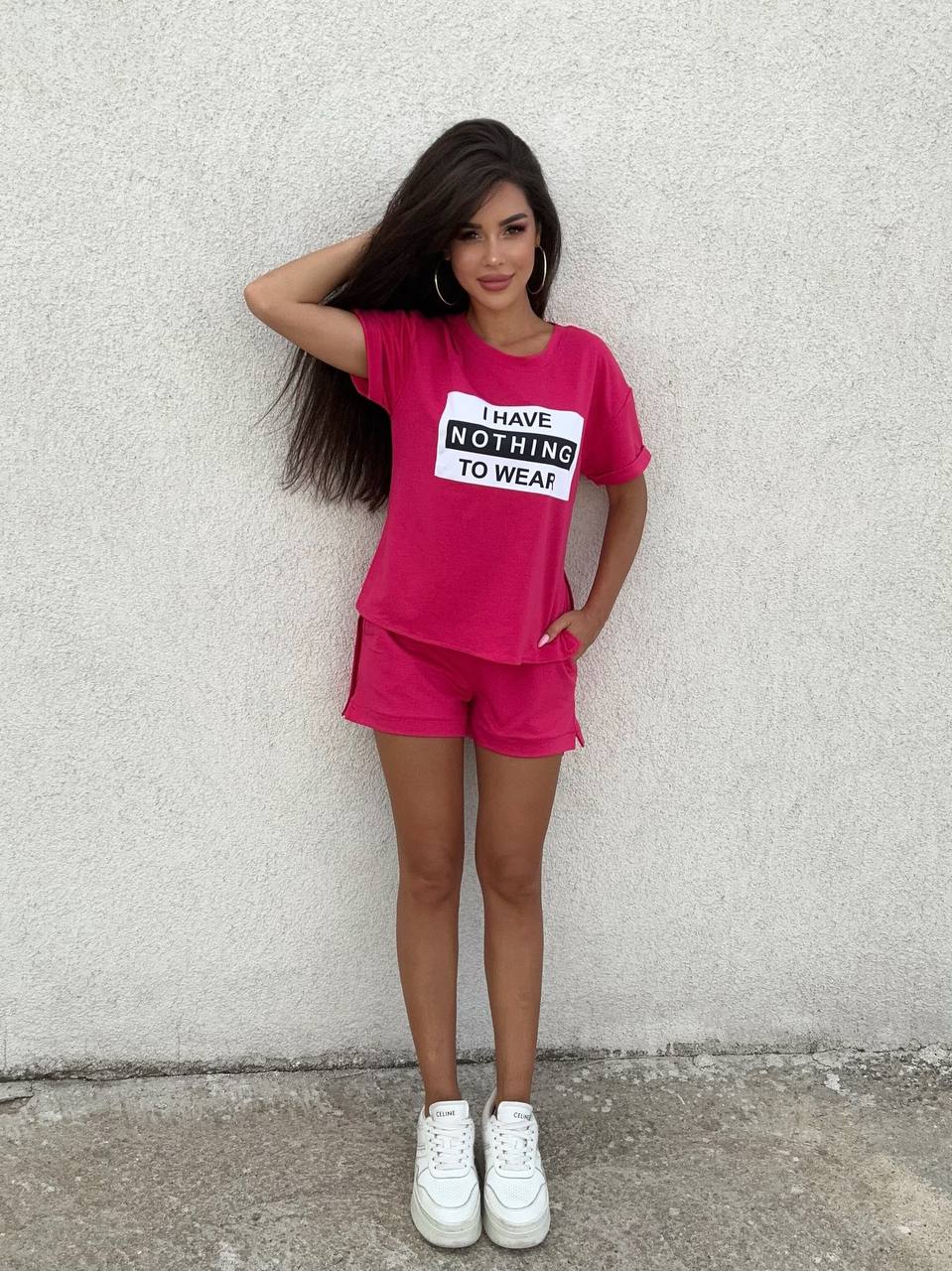 Woman wearing a pink outfit with a t-shirt and shorts, standing against a plain wall.