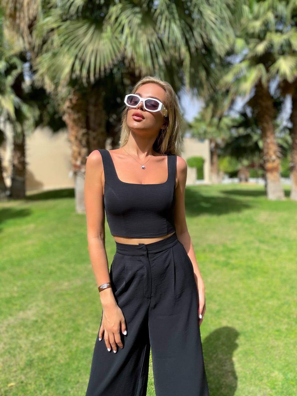 Woman in black outfit with sunglasses standing in a park with palm trees.