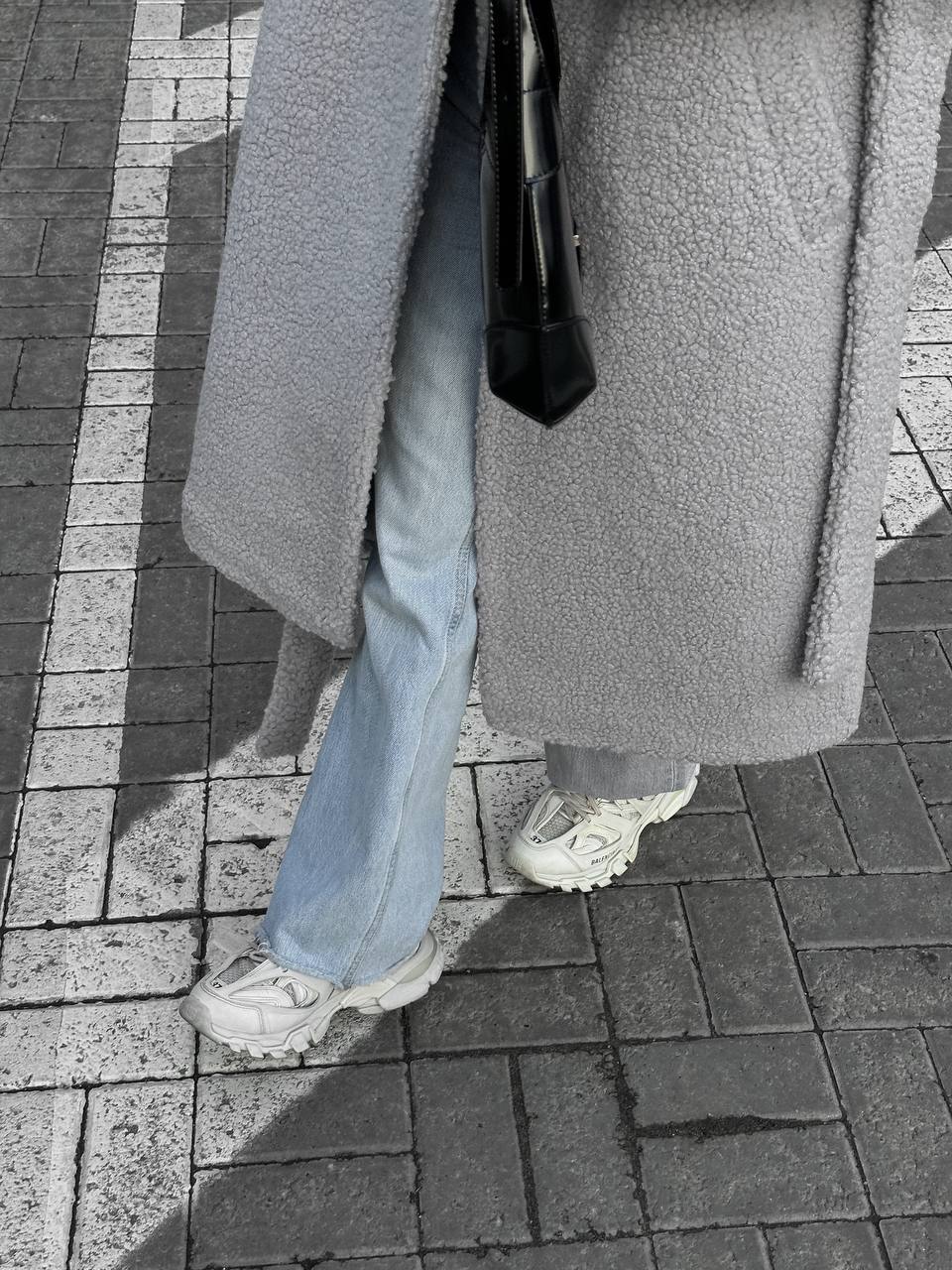 Person wearing a long gray coat, light blue jeans, and white sneakers on a sidewalk.