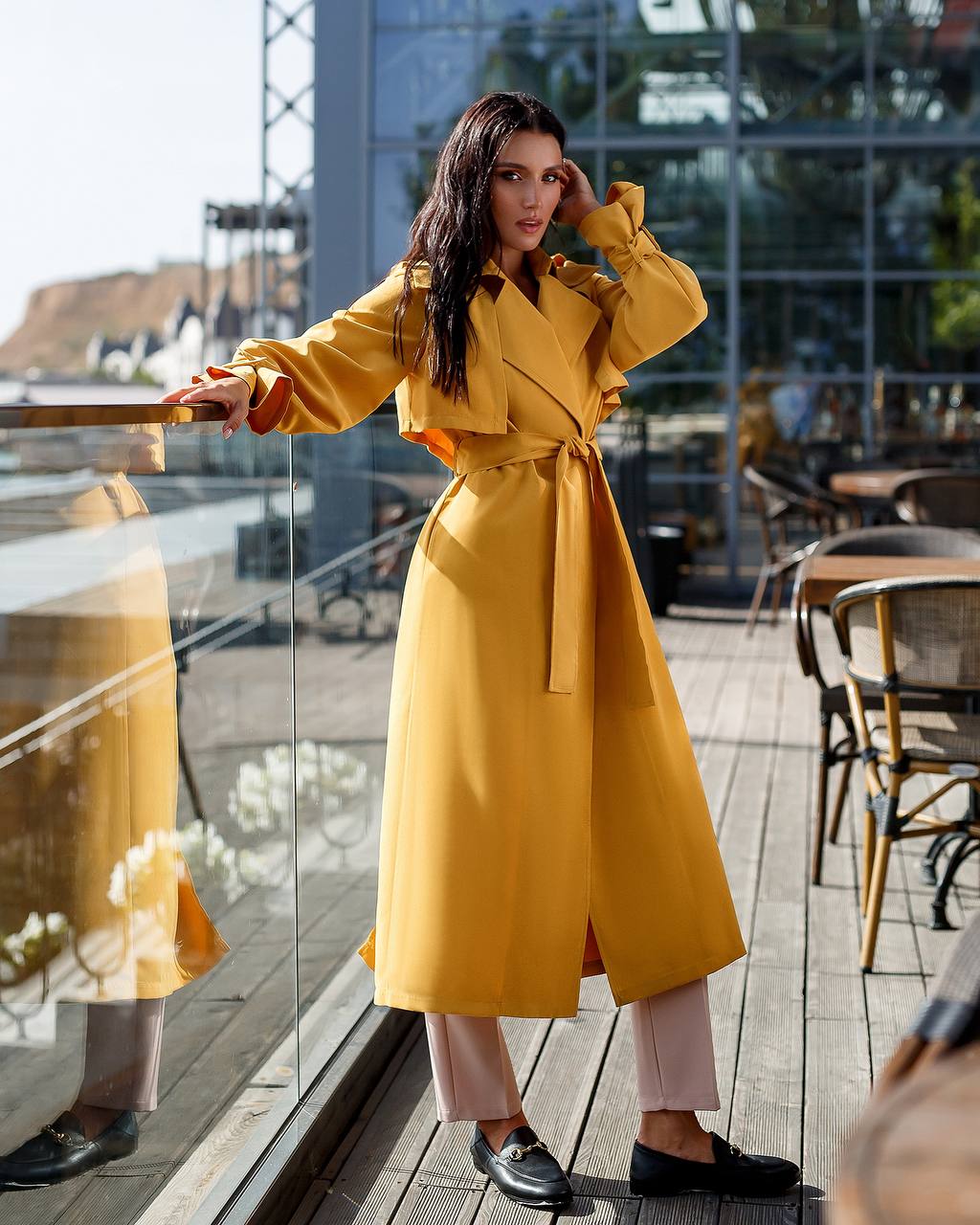 Woman in a yellow trench coat standing on a wooden deck with tables and chairs.