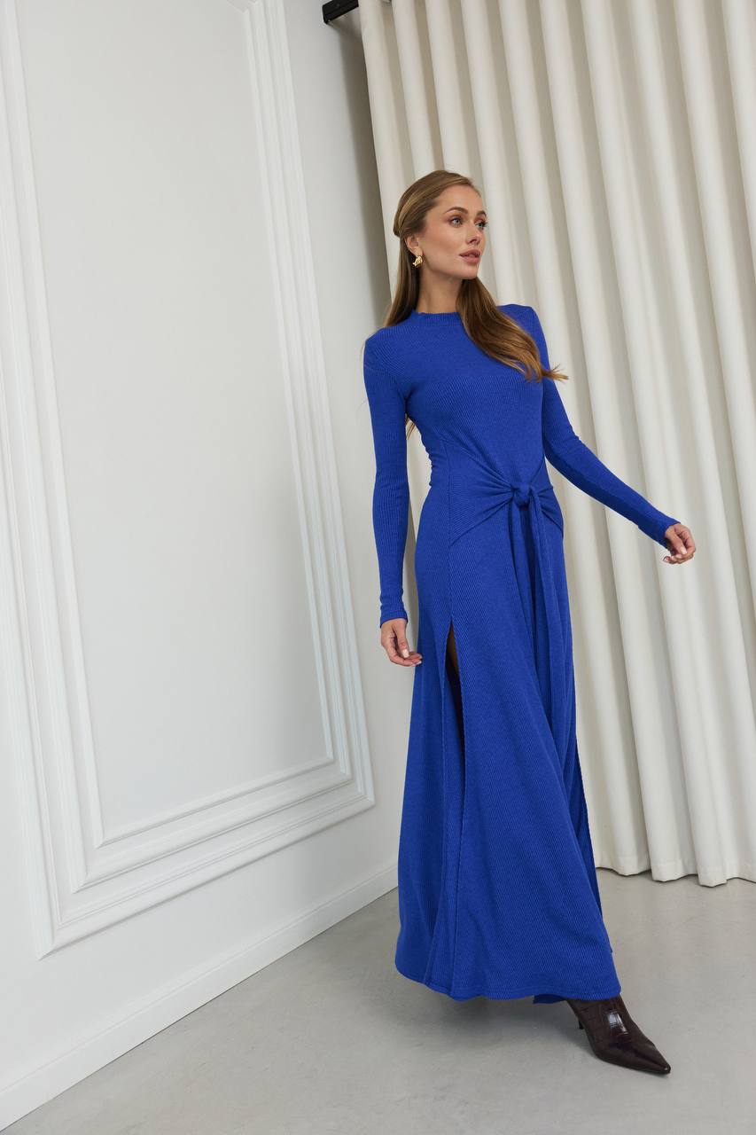 Woman wearing a blue dress standing against a white wall with curtains.